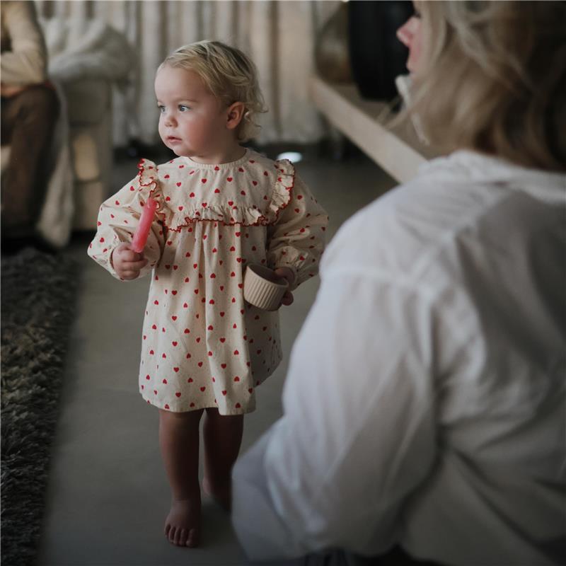 Toddler in a heart-patterned dress holding a pink spoon and a small beige container, walking in a cozy setting.