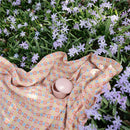 Mushie silicone baby food container in pink, placed on colorful fabric among light purple flowers.