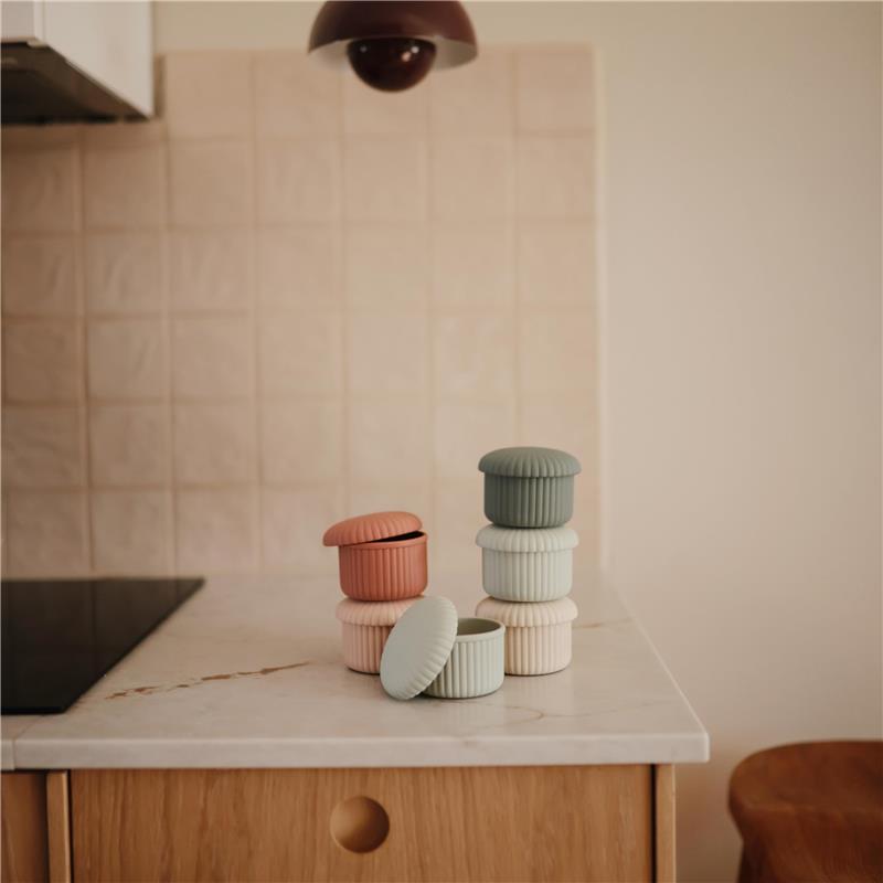 Mushie Silicone Baby Food Containers in pink, green, and beige stacked on a marble countertop in a kitchen setting.