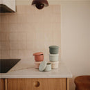 Mushie Silicone Baby Food Containers in pink, green, and beige stacked on a marble countertop in a kitchen setting.