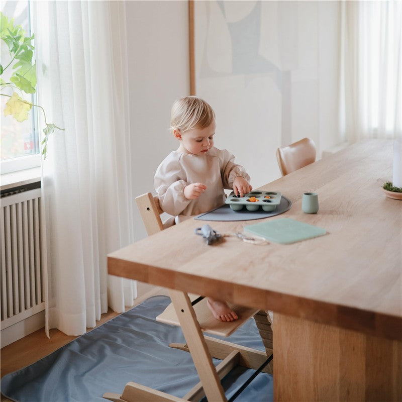 A toddler in a light-colored outfit uses a Mushie Baby Food Freezer Tray on a wooden table, surrounded by a bright, cozy kitchen.