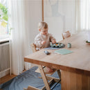 A toddler in a light-colored outfit uses a Mushie Baby Food Freezer Tray on a wooden table, surrounded by a bright, cozy kitchen.