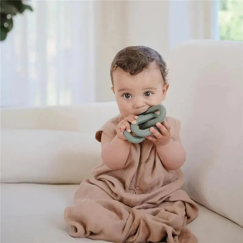 Baby sitting on a couch, holding a green Mushie silicone teether toy, wearing a light brown romper.