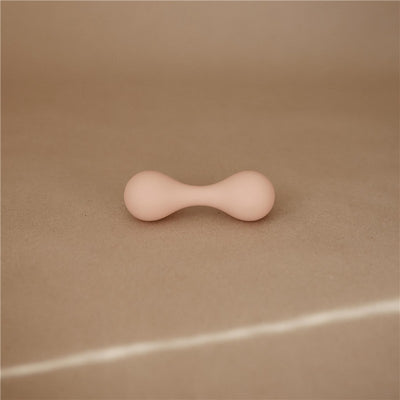 Silicone baby rattle toy in blush color, shaped like a dumbbell, resting on a light brown background.