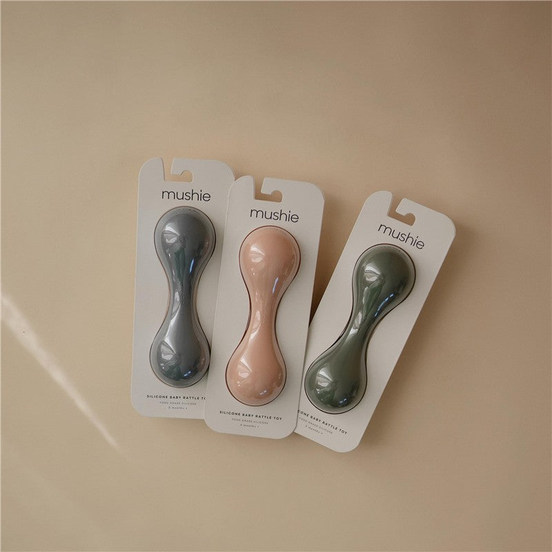 Three Mushie silicone baby rattle toys in gray, peach, and green, displayed on packaging, with the brand name visible.