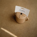 Mushie Silicone Snack Cup in natural color with a lid and two handles, displayed on a brown surface.