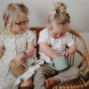 Two children sitting on a wicker chair, each holding a Mushie Silicone Snack Cup; one in brown and the other in green, engaged in playful activity.
