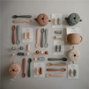Mushie Silicone Snack Cups and spoons in various colors on a neutral surface, showcasing toddler feeding products and accessories.