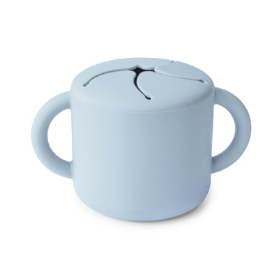 Mushie silicone snack cup in powder blue with two handles and a lid featuring slits for easy access to snacks.