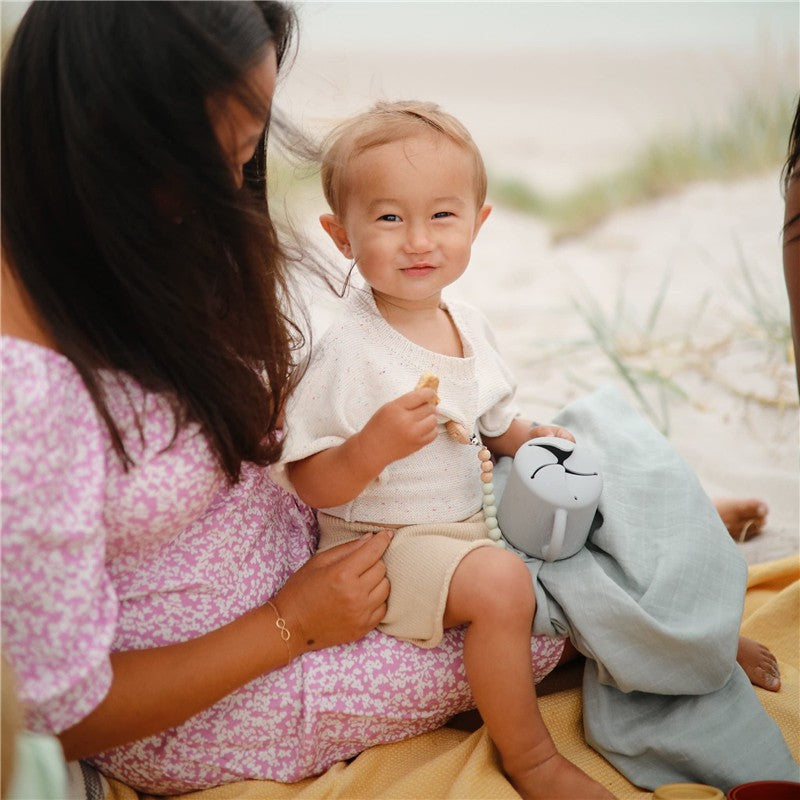 A smiling child holds a snack and a powder blue Mushie silicone snack cup while sitting on a blanket with an adult.