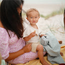 A smiling child holds a snack and a powder blue Mushie silicone snack cup while sitting on a blanket with an adult.