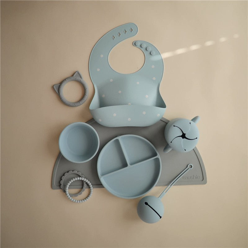 Mushie Silicone Snack Cup in Powder Blue surrounded by a bib, various bowls, and a teething toy on a neutral background.