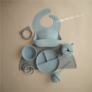 Mushie Silicone Snack Cup in Powder Blue surrounded by a bib, various bowls, and a teething toy on a neutral background.