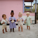 Four children stand on sand, each holding a Mushie silicone snack cup in varying colors, with a pink beach house in the background.