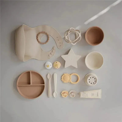 Silicone baby suction bowl in ivory, surrounded by feeding accessories like spoons, teethers, and bib.