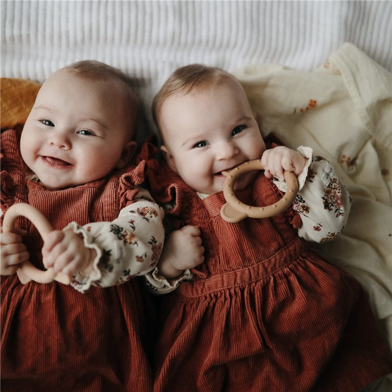 Twins smile while holding a Mushie silicone bear teether in a cozy setting, wearing matching rust-colored dresses.