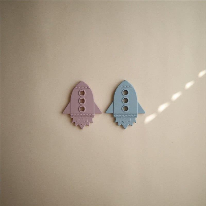 Silicone rocket-shaped teethers in pink and blue colors, designed for infants, placed on a light-colored surface.