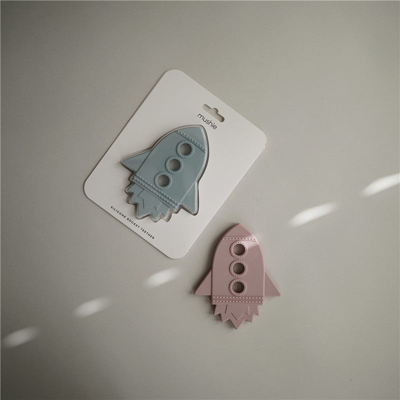 Mushie Silicone Rocket Teether in cloud blue color displayed on packaging next to a pink version, featuring multiple holes for grip.