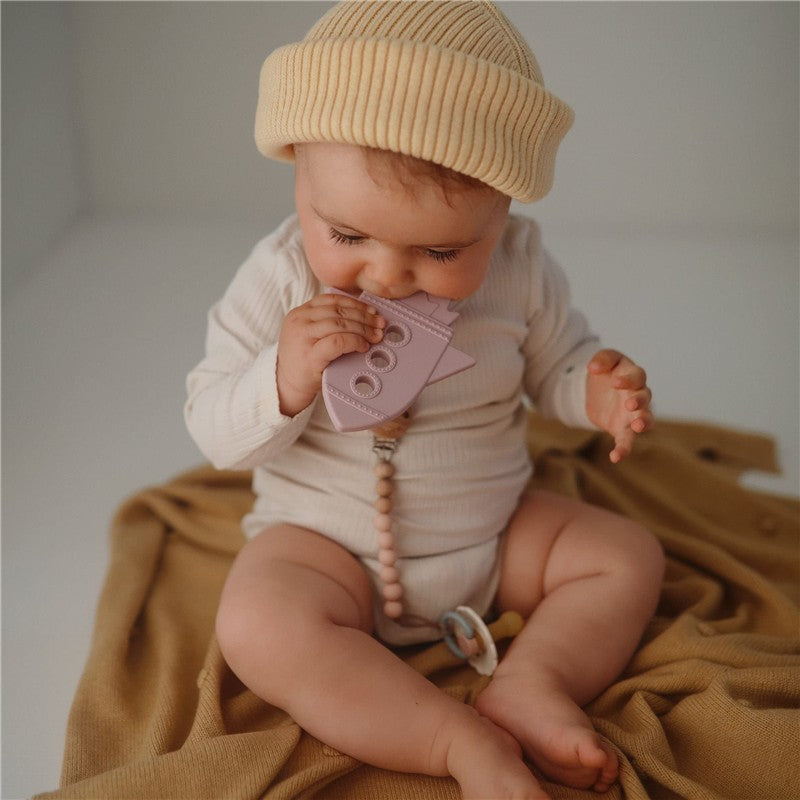 Baby sitting on a soft fabric, chewing on a lilac silicone rocket teether, wearing a cream-colored hat and light outfit.