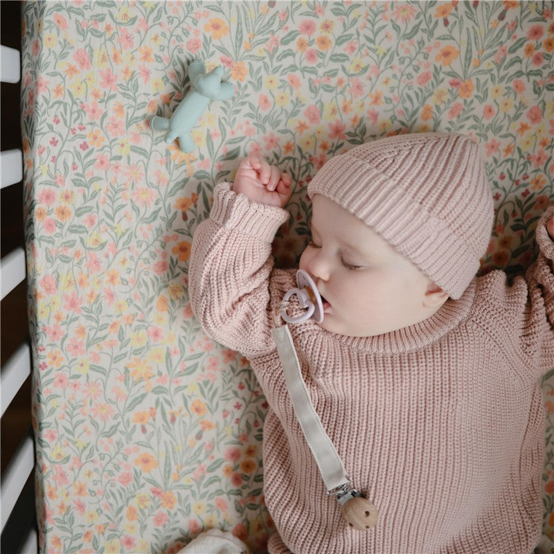 Baby dressed in a pink knitted sweater and hat, sleeping on a floral blanket next to a blue Mushie dog figurine teether.