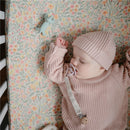 Baby dressed in a pink knitted sweater and hat, sleeping on a floral blanket next to a blue Mushie dog figurine teether.