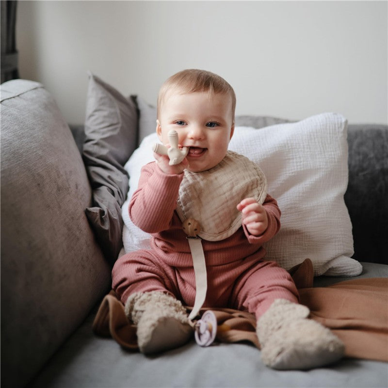 Baby smiling while holding a Mushie Silicone Lamb Figurine Teether, wearing a pink outfit and a beige bib, sitting on a couch.