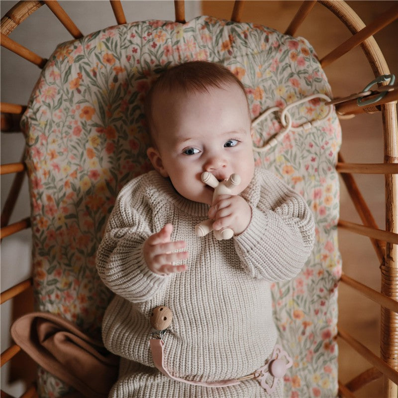 A baby in a cozy, knitted outfit holds a Mushie silicone lamb figurine teether while sitting in a floral-patterned chair.
