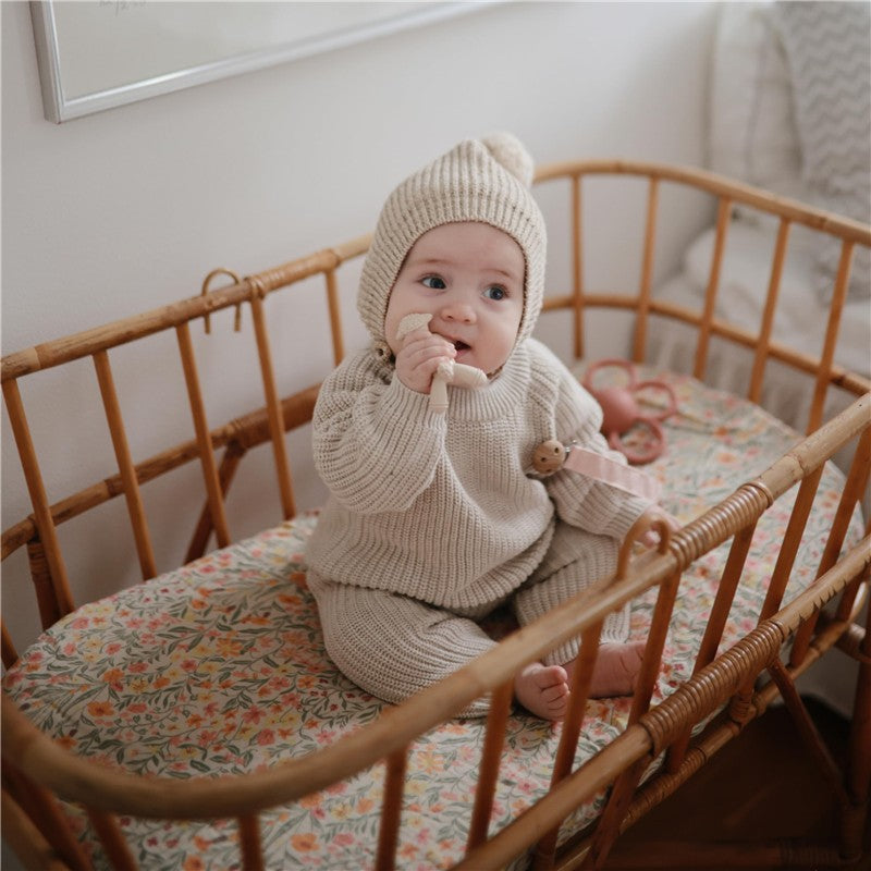 Baby sitting in a woven crib, wearing a beige knit outfit and hat, holding a teether.