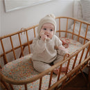 Baby sitting in a woven crib, wearing a beige knit outfit and hat, holding a teether.
