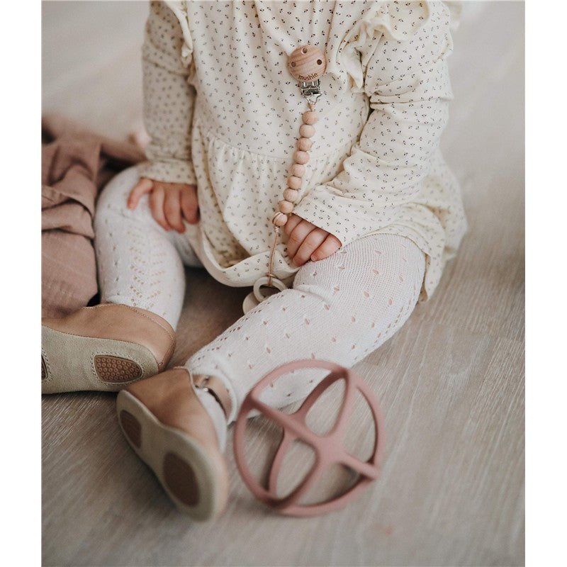 Mushie Silicone Ball Teether in Woodchuck color next to a child wearing a patterned dress and shoes, with a pacifier clip visible.