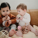 Two babies sit on a couch, one holding a stack of colorful silicone cups and the other chewing on a silicone teether, with a pink flower design.