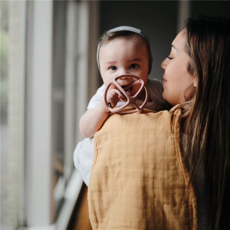 Baby holding a pink Mushie Silicone Ball Teether while being cuddled by a woman, with soft natural lighting in the background.