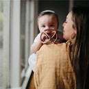 Baby holding a pink Mushie Silicone Ball Teether while being cuddled by a woman, with soft natural lighting in the background.