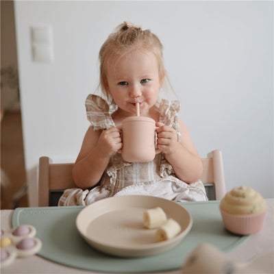 A child with blonde hair holds a blush pink silicone training cup with a straw, sitting at a table with fruit and a cupcake.