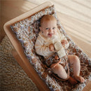 Infant smiling while holding a silicone bottle with decorative floral design in a cozy bouncer.