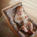 Baby in a floral-patterned seat holding a silicone baby bottle with a natural and shifting sand design.
