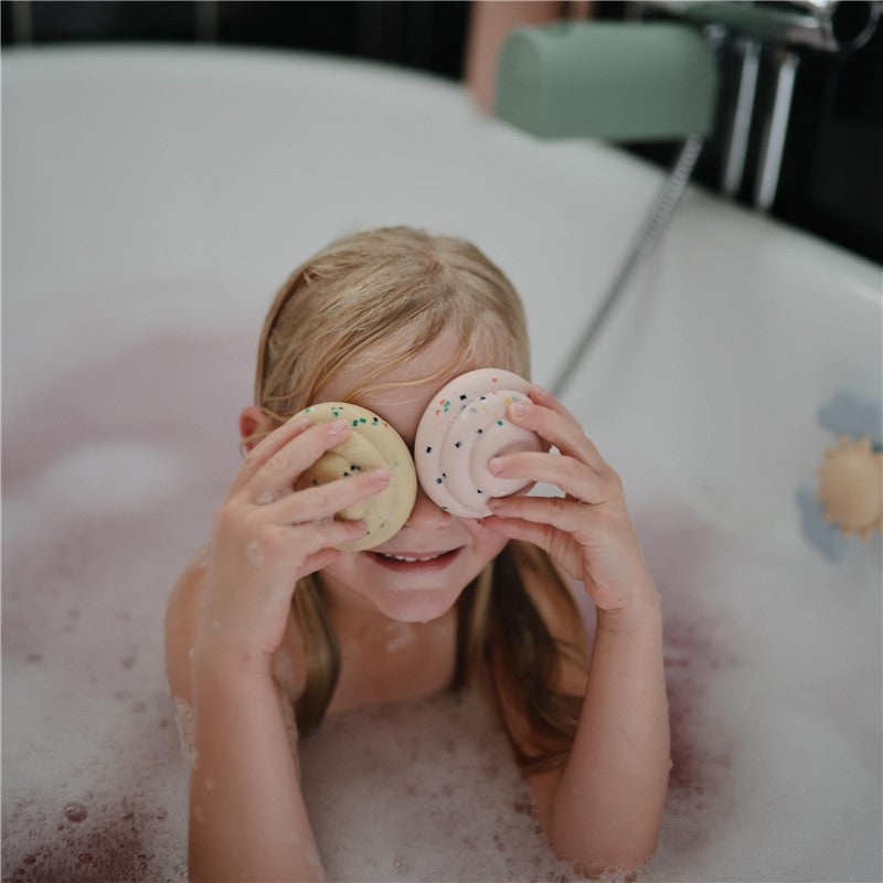 A smiling child in a bathtub holds a pink and yellow cupcake bath toy to her face, surrounded by bubbles.