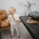 A toddler in a white knitted outfit stands next to a chair, holding a yellow and beige silicone teething ring.
