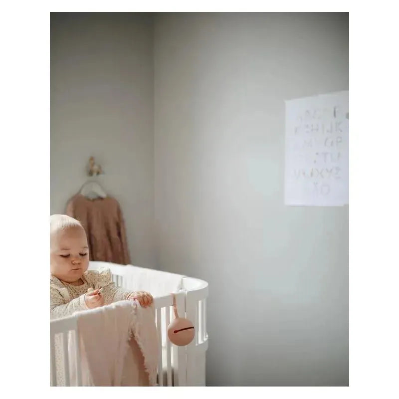 Baby in a crib holds a spoon, with a blush-colored Mushie silicone pacifier holder hanging nearby.
