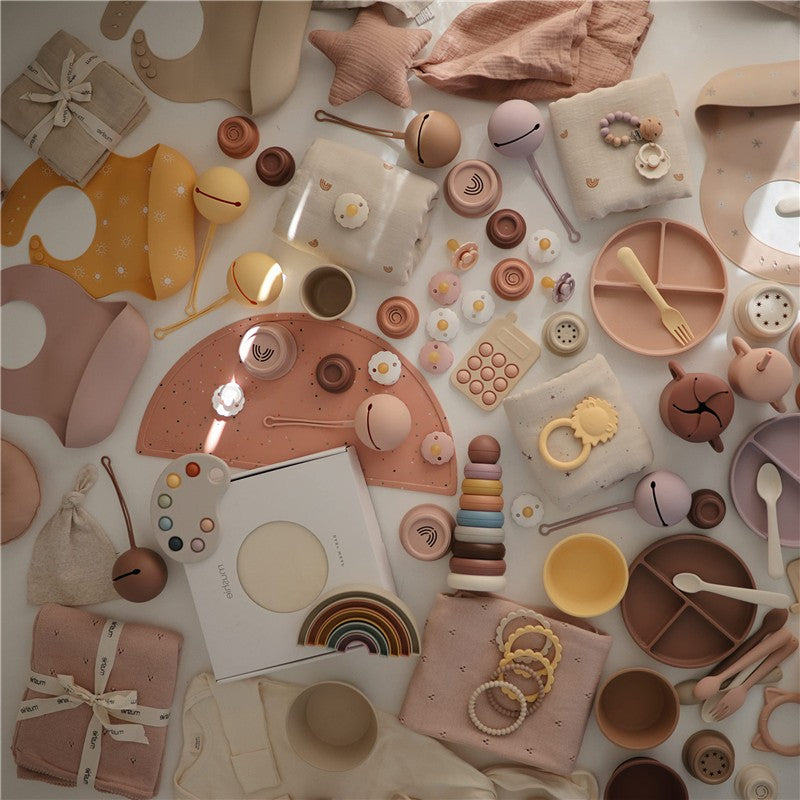Mushie Silicone Pacifier Holder Case in blush color amidst various baby accessories and tableware, featuring soft pastel tones and playful designs.