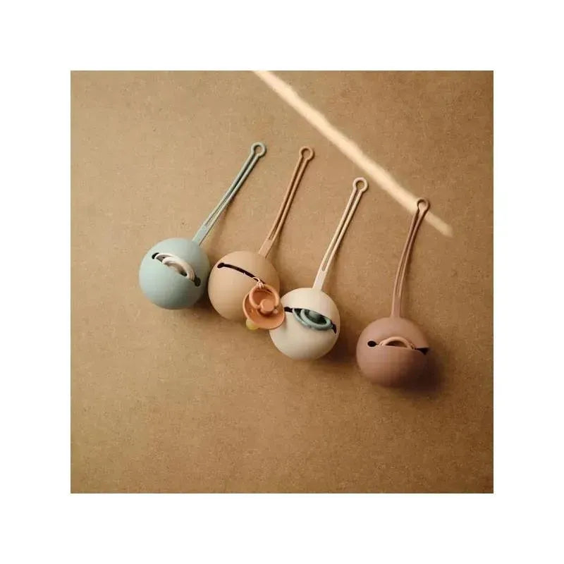 Mushie Silicone Pacifier Holder Cases in various colors, designed to store and protect pacifiers, arranged on a brown surface.