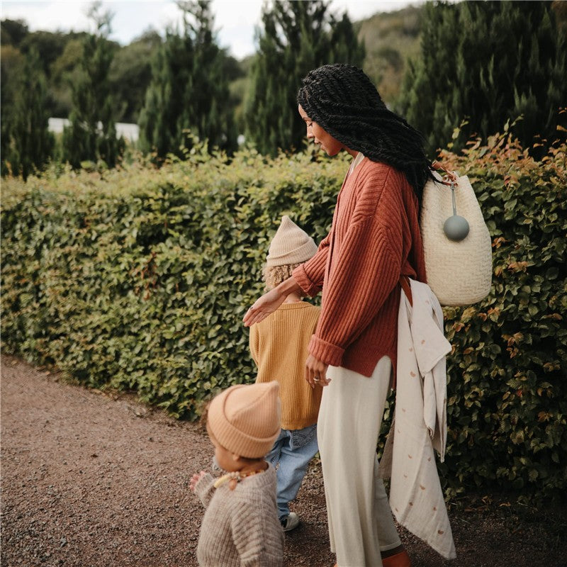 Mushie Silicone Pacifier Holder Case in Dried Thyme featured in a stylish outdoor family scene with a mother and two children.