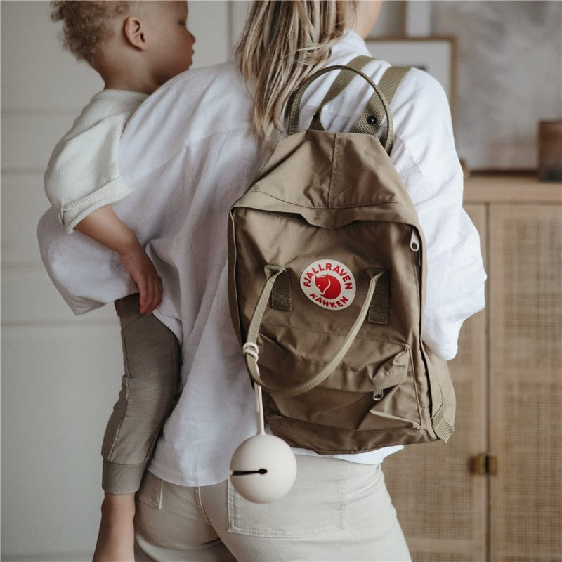 Mushie Silicone Pacifier Holder Case in Powder Blue attached to a Khaki Fjällräven Kånken backpack being carried by a woman holding a child.