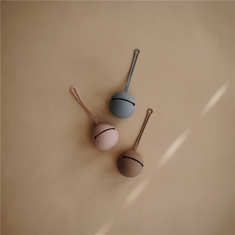 Silicone pacifier holder cases in stone, pink, and blue colors, laid against a beige background.