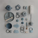Mushie Silicone Pacifier Holder Case in stone color, surrounded by various baby accessories including toys and feeding items.