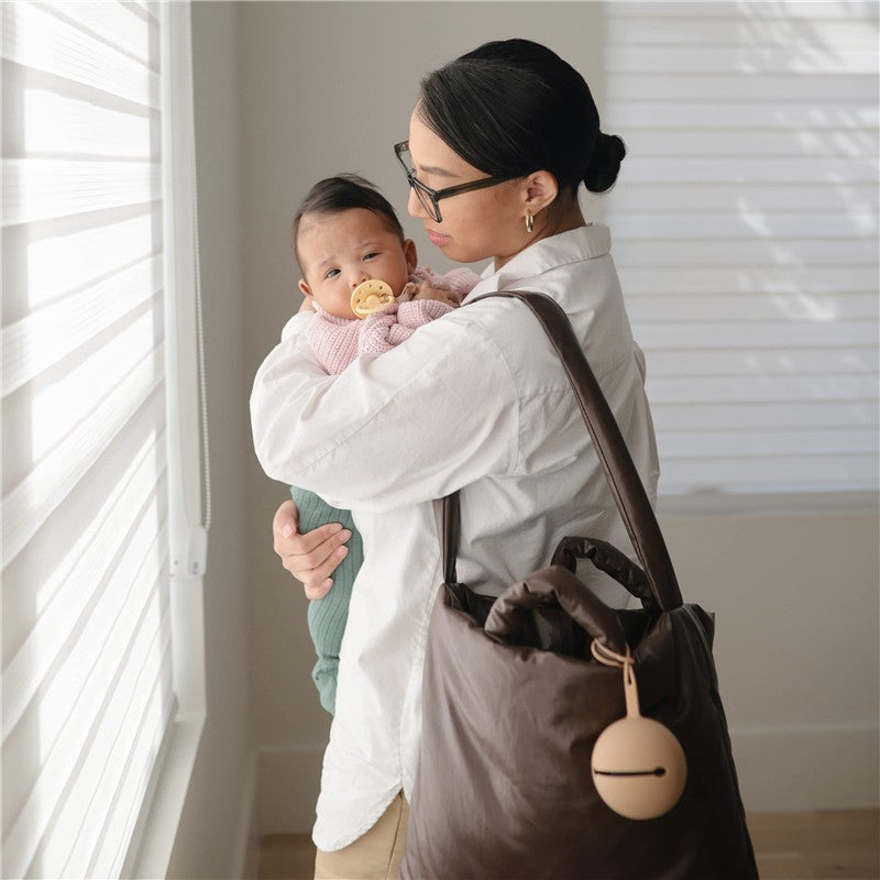 A woman in a white shirt holds a baby with a pacifier, next to a brown bag with a silicone pacifier holder.