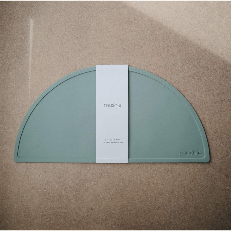 Mushie silicone placemat in Cambridge Blue, featuring a rounded design with a white label detailing care instructions.