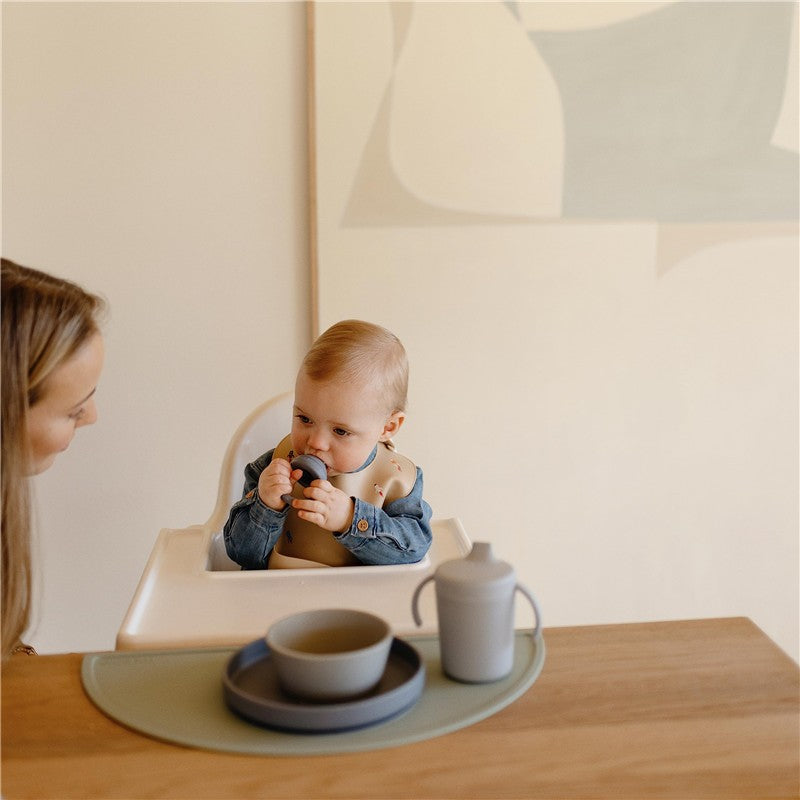 Mushie Silicone Placemat in Cambridge Blue with a child in a high chair, holding a toy, and dishes on the table.