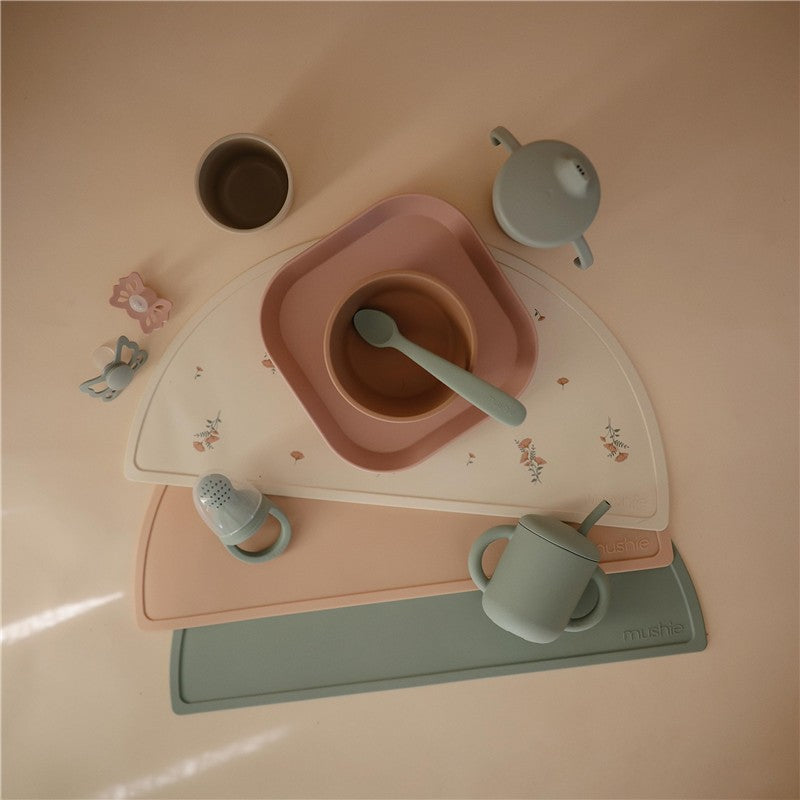 Mushie Silicone Placemat in Cambridge Blue with pink and beige bowls, utensils, and a cup, arranged on a light surface.