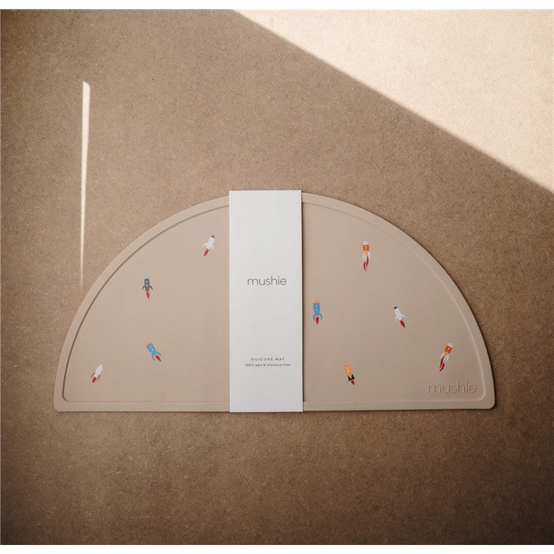 Mushie Silicone Placemat in a rocket ship design, featuring colorful rocket graphics on a beige surface.
